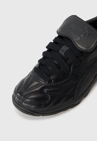 Sort lædersneaker med textureret overflade, snørebåndsdesign og velcro-strop. Har Puma-branding og rund tå.