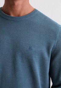 Sweter dziany w odcieniu niebieskiego teal, z prążkowanym okrągłym dekoltem oraz subtelnym wzorem teksturalnym. Małe logo na klatce piersiowej. Miękka tkanina.