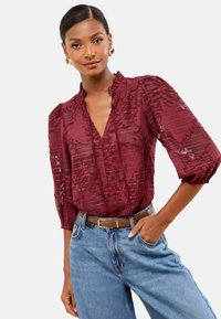 Femme portant un chemisier en burgundy transparent texturé avec des manches 3/4, un jean bleu clair, une ceinture marron et des boucles d'oreilles en or en forme de créole, posant avec la main dans la poche.