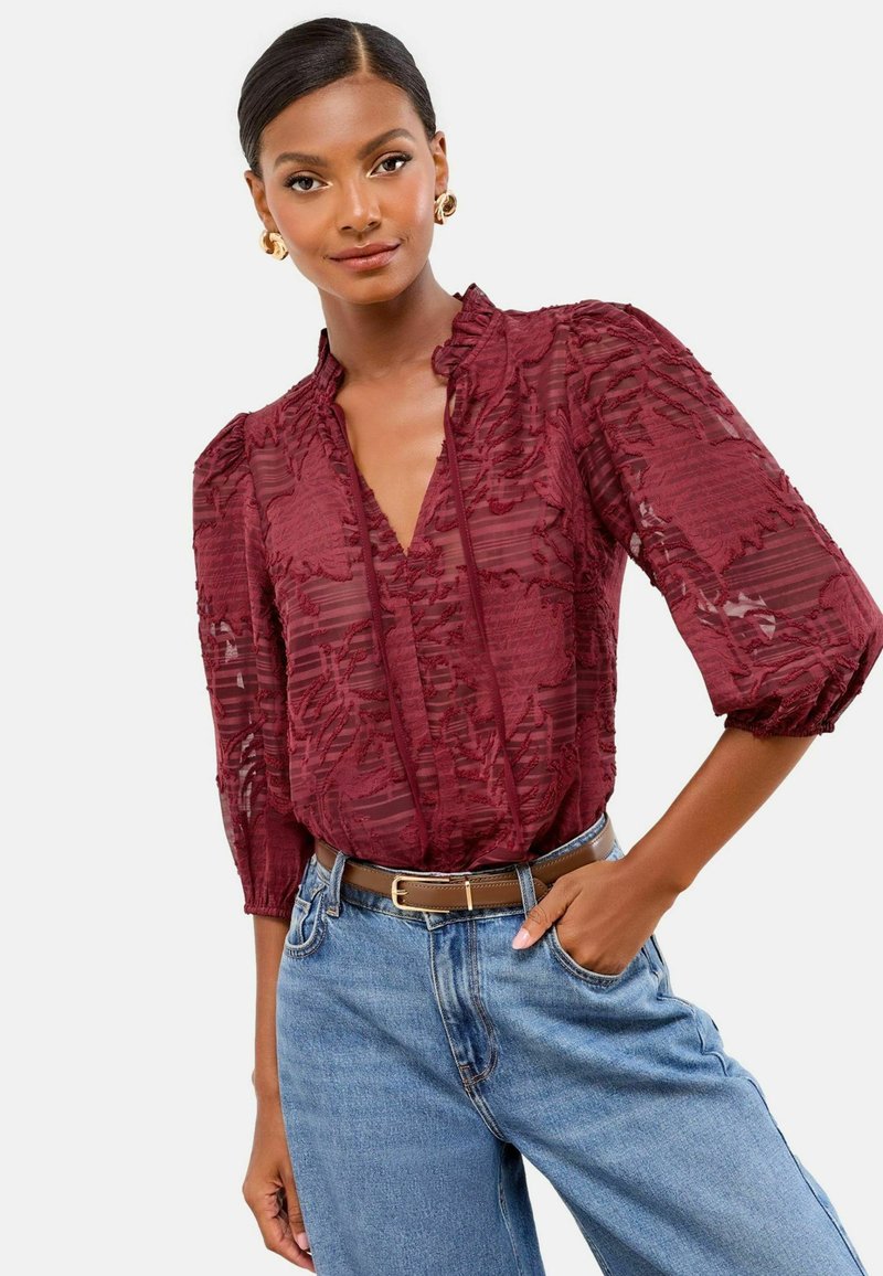 Femme portant un chemisier en burgundy transparent texturé avec des manches 3/4, un jean bleu clair, une ceinture marron et des boucles d'oreilles en or en forme de créole, posant avec la main dans la poche.