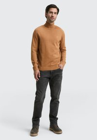 Maglione a collo alto marrone chiaro, con polsini e orlo a coste, abbinato a jeans grigi scuri e stivali marroni con lacci. Modello in piedi con le mani nelle tasche.