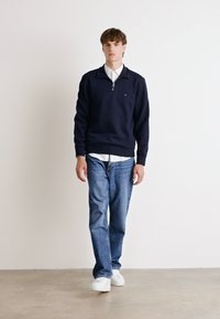 Navy zip-neck sweatshirt paired with a white collared shirt, light blue jeans, and white sneakers. Smooth fabric; relaxed fit.
