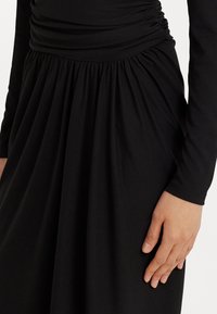 Vestido negro de manga larga con cintura fruncida y falda recogida, con un primer plano de una mano descansando en la cadera.