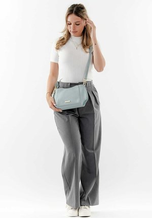 Bolso de mano de cuero texturizado en azul claro, con cierre de cremallera y herrajes en dorado, sostenido por una persona que lleva pantalones grises y una blusa blanca ajustada.