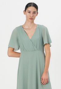 Ljusgrön klänning med v-ringning och korta volangärmar. Tillverkad av ett slätt tyg, med draperad design och subtil rörelse.