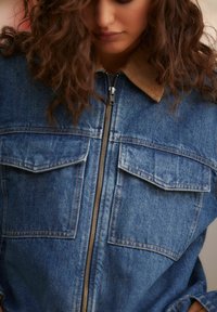 Denim-takki, jossa on vetoketju, kaksi rintataskua ja korduroitukaulus. Vaalea sininen kangas, jossa on hieman teksturoitu pinta.