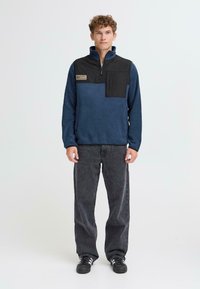 Sudadera de forro polar en azul marino y negro, con cuello de media cremallera y un bolsillo en el pecho. Llevada con pantalones de mezclilla negros y zapatillas.