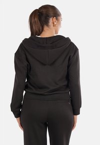 Felpa nera con cappuccio, realizzata in tessuto morbido, caratterizzata da un cappuccio ampio, una vestibilità comoda e un bordo a costine. La vista posteriore mostra una texture liscia e un design pulito.
