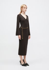 Pruun soonikko dress, millel on V-kaelus, pikad flared varrukad, kuldne ketist vöö ja mustad terava ninaga kontsad. Sile tekstuur, istuv siluett.