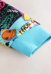Poignet de manche en maille côtelée bleu clair avec des imprimés colorés mettant en avant un personnage orange de dessin animé, un texte en gras et des motifs style graffiti.