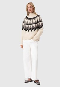 Πουλόβερ από μαλλί με ψηλό γιακά, με γεωμετρικό σχέδιο σε μαύρο, γκρι και κρέμ χρώμα; συνδυασμένο με φαρδιά άσπρα παντελόνια και γκρι παντόφλες.