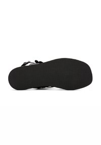 Sandalia negra con suela de goma texturizada, diseño abierto y correas ajustables. Presenta una suela plana y elementos de estilo minimalista.