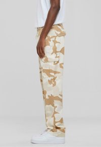 Cargo pants in beige camouflage pattern, cotton fabric, side pockets, relaxed fit, paired with white sneakers.