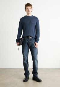 Maglione a coste navy, jeans blu scuro e stivaletti neri alla caviglia. La modella tiene una piccola borsa nera ed è in piedi su un pavimento di cemento.