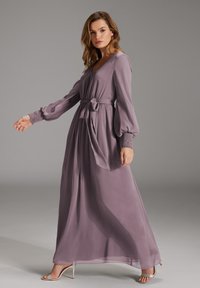 Langes, fließendes Maxikleid aus weichem Lavendeltuch. Es hat einen tiefen V-Ausschnitt, eine taillierte Taille und geraffte Ärmelbündchen. Kombiniert mit Schnürsandalen.