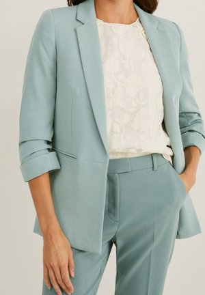 Femme portant un blazer bleu clair aux manches retroussées et un pantalon assorti, associé à un chemisier blanc à motif floral.