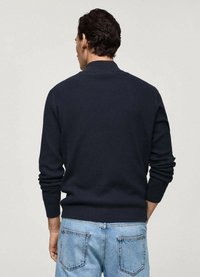 Navy ribbed knit sweater with a high collar, fitted cuffs, and a relaxed back design, paired with light blue denim jeans.