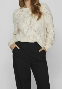 Sweter dzianinowy w off-white z diamentowym wzorem, otwartym splotem i długimi rękawami, zestawiony z czarnymi spodniami fasonu tailored z kieszeniami bocznymi.