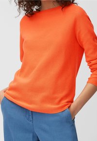 Pull tricoté orange vif avec un col rond et des manches longues. La texture est lisse et extensible, associé à un pantalon bleu.