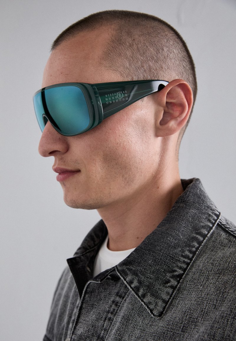 Grüngetönte Sonnenbrille mit einem glatten Kunststoffrahmen, verspiegelten Gläsern und kantigem Design; verfügt über numerische Markierungen an den Bügeln.