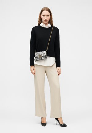 Femme portant un pull noir sur une chemise blanche, un pantalon ample beige, des talons noirs et un sac à bandoulière à motif peau de serpent sur un fond uni.