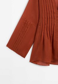 Rustfarvet bluse med plisseret design på ærmerne og kroppen. Lavet af letvægtsstof og har en løs, flydende silhuet.