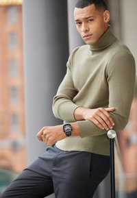 Un pull à col roulé côtelé vert olive associé à un pantalon noir, arborant une montre moderne avec un bracelet noir et des accents en argent.