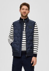 Die marineblaue gesteppte Weste verfügt über einen hohen Kragen und einen Reißverschluss und wird über einem weiß-blauen gestreiften Pullover getragen. Das Material sieht glatt und gepolstert aus.
