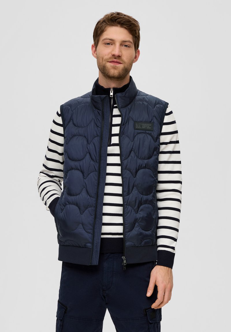 Die marineblaue gesteppte Weste verfügt über einen hohen Kragen und einen Reißverschluss und wird über einem weiß-blauen gestreiften Pullover getragen. Das Material sieht glatt und gepolstert aus.