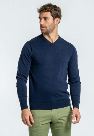 Pull col V bleu marine en tricot avec des manches longues et des poignets côtelés, associé à un pantalon vert olive.