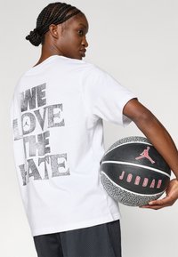 Balta trumpa rankovėmis marškinėliai su dideliu juodu blizgučiu tekstu "MUM PATINKA NEAPYKANTA" ant nugaros. Juoda ir pilka tekstūruota krepšinio kamuolys su raudonais akcentais.