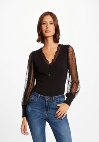 Body nero a maniche lunghe con scollatura a V profonda, con rifinitura in pizzo e maniche a sbuffo trasparenti con pois. Abbinato a jeans blu.