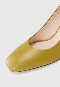 Zielony skórzany but na niskim obcasie z kwadratowym noskiem i jasnoróżową podszewką wnętrza na białym tle.