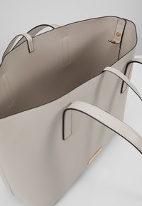 Beige læder tote taske med sorte kanter, der har to håndtag og et guldaccent på fronten. Indersiden er rummelig og uden foring.