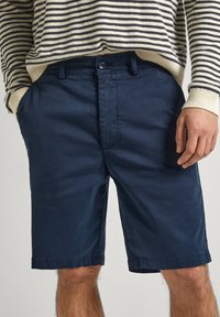 Shorts bleu marine en coton lisse, avec fermeture à bouton, poches avant et ourlet légèrement cintré. Pull rayé visible au-dessus.