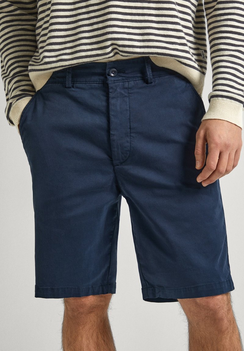 Shorts bleu marine en coton lisse, avec fermeture à bouton, poches avant et ourlet légèrement cintré. Pull rayé visible au-dessus.