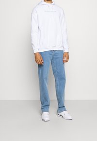Felpa bianca con cappuccio, dotata di tasche frontali e testo azzurro, abbinata a jeans azzurri e sneakers bianche. Design semplice e casual.