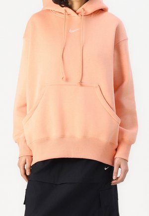 Personne portant un sweat à capuche couleur pêche avec une poche avant et un petit logo blanc Nike, associé à une jupe noire avec un petit logo blanc Nike.