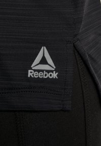 Black athletic shirt featuring a textured fabric, side slit, and a reflective grey Reebok logo on the hem.