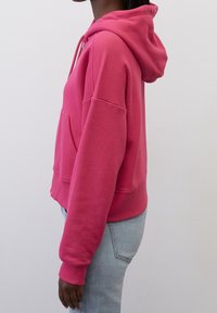 Vista lateral de una persona que lleva puesto un sudadera rosa brillante y jeans azul claro frente a un fondo blanco liso.