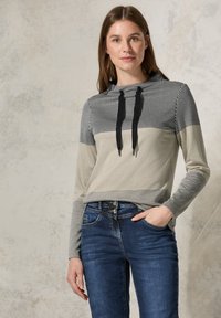 Gestreepte trui in zwart, wit en beige, met een capuchon en zwarte trekkoorden. Gepaard met donkerblauwe jeans.