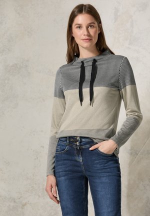 Maglione a righe nei toni del nero, bianco e beige, con cappuccio e lacci neri. Abbinato a jeans blu scuro.