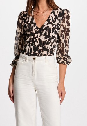 Vrouw draagt een zwart-beige dierenprint V-hals blouse met pofmouwen en wit hoogtaillepantalon, geaccentueerd met gelaagde gouden kettingen.