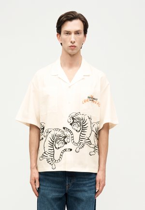 Άνδρας που φοράει κοντομάνικο κρεμ πουκάμισο με μαύρες απεικονίσεις τίγρης και πορτοκαλί κείμενο στο στήθος, συνδυασμένο με μπλε τζιν, στέκεται μπροστά σε λευκό φόντο.