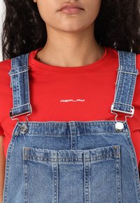 Camiseta vermelha com o logótipo "REPLAY" acima de um macacão de denim. O macacão apresenta alças ajustáveis, acessórios em prata e um bolso frontal com costuras verticais.