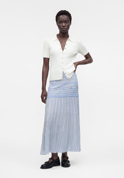 Γυναίκα στέκεται με το χέρι στη μέση φορώντας λευκό πουκάμισο με κοντά μανίκια και κουμπιά, μακρύ μπλε φούστα με σχέδιο και μαύρα παπούτσια με αγκράφα σε λευκό φόντο.