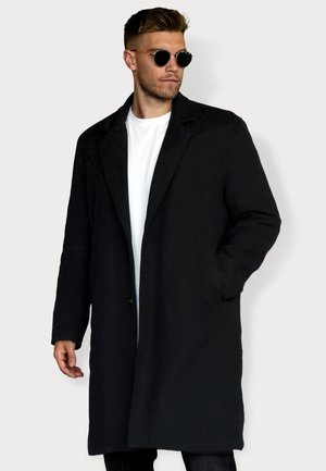 Schwarzer langer Mantel mit taillierter Passform, ausgestattet mit einem Reverskragen, einem einzelnen Knopfverschluss und seitlichen Taschen. Über einem weißen T-Shirt getragen.