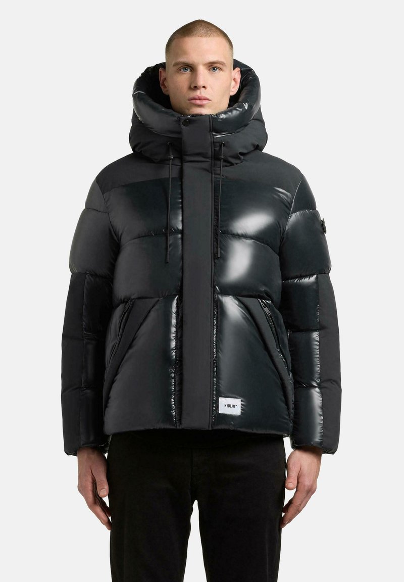 Pufferjack in glanzend zwart materiaal, met een hoge kraag, ritszakken en een label aan de onderrand. Kenmerkend doorgestikt ontwerp.