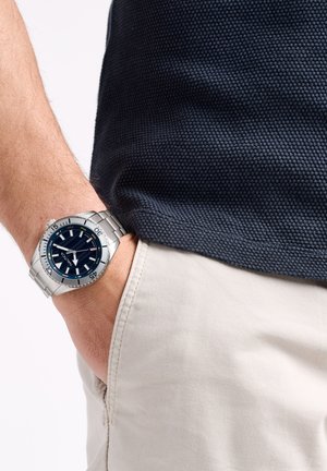 Reloj de acero inoxidable plateado con una esfera de color azul marino, que presenta marcadores de hora blancos, una pantalla de fecha y una pulsera de eslabones metálicos.