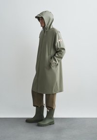 Wodoodporny płaszcz w kolorze oliwkowej zieleni z kapturem, przednimi kieszeniami i logo marki na rękawie, zestawiony z brązowymi spodniami i zielonymi gumowymi butami.